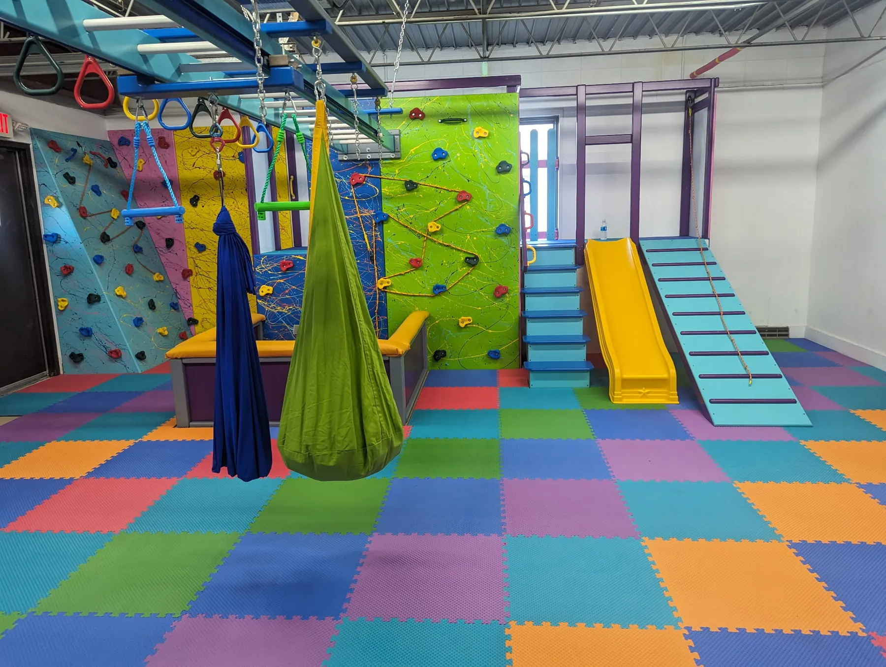 Large sensory gym with climbing walls, a cocoon swing, a ball pit, stairs, a yellow slide, and a slatted ramp on checkered foam tiles