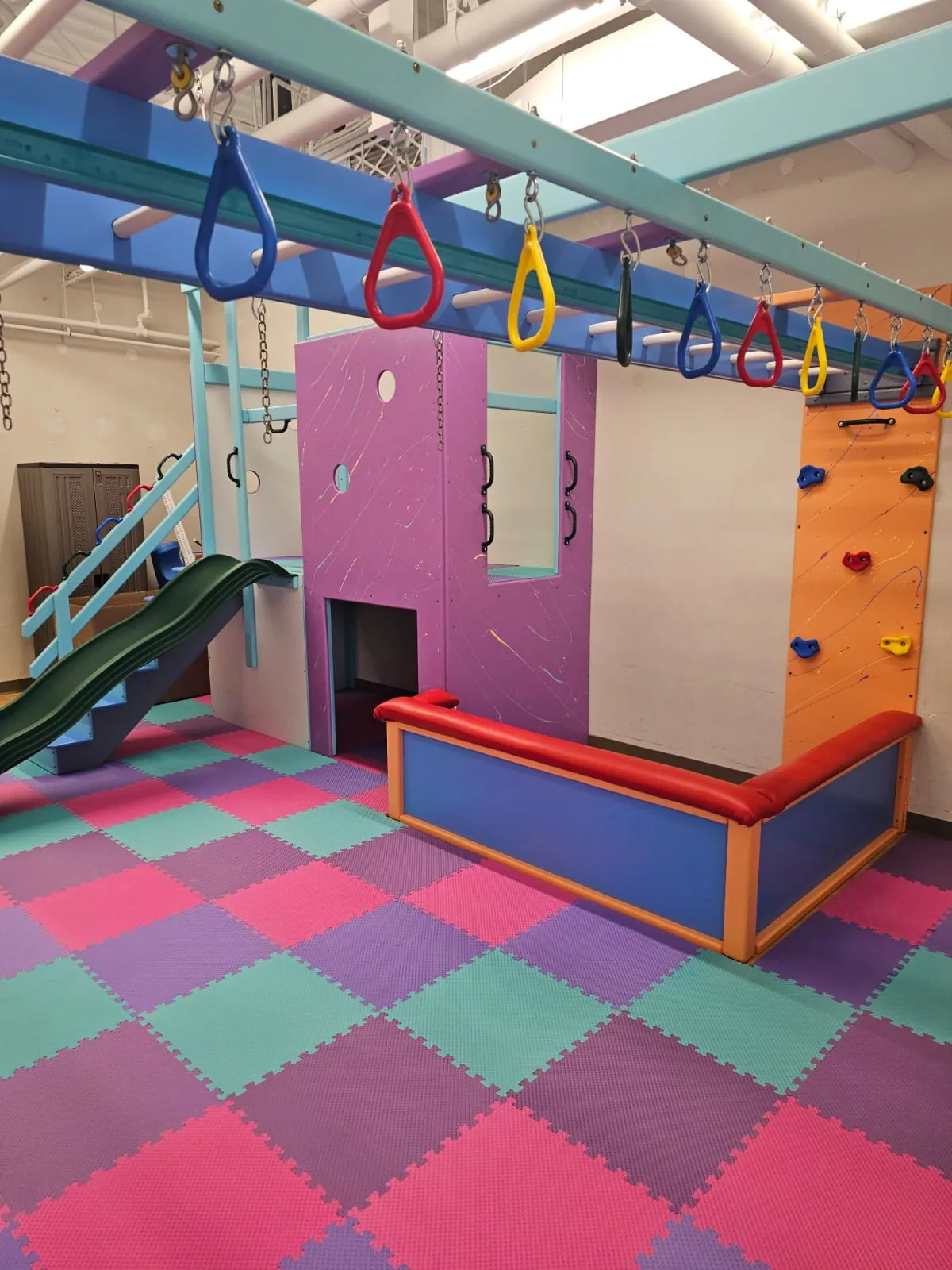 Wide view of a purple playhouse, green slide, overhead ring track, and orange climbing wall above pink and teal foam tiles