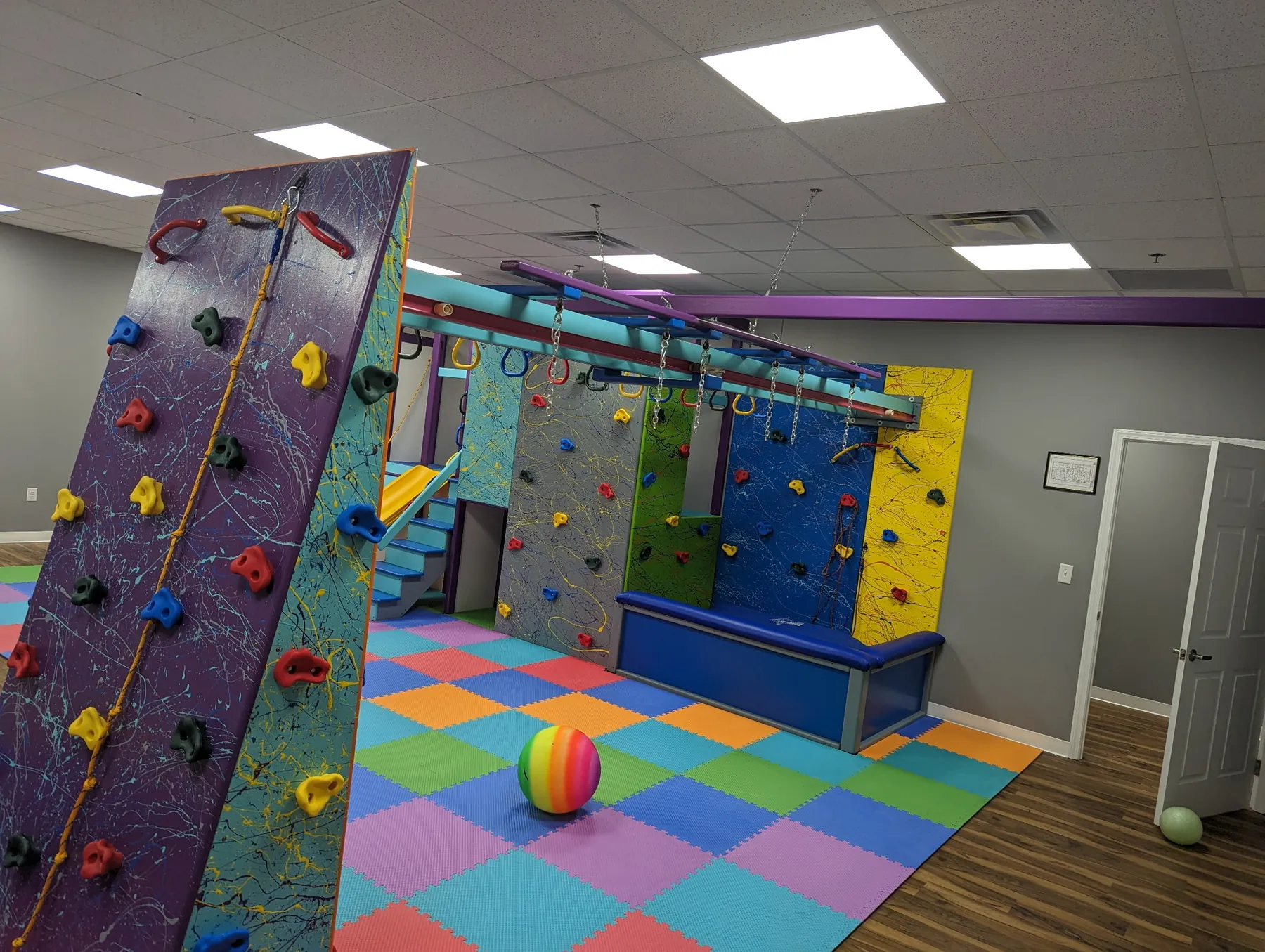 Purple angled climbing wall in front of climbing panels, a padded bench, and a ring track in a sensory gym with checkered foam tiles