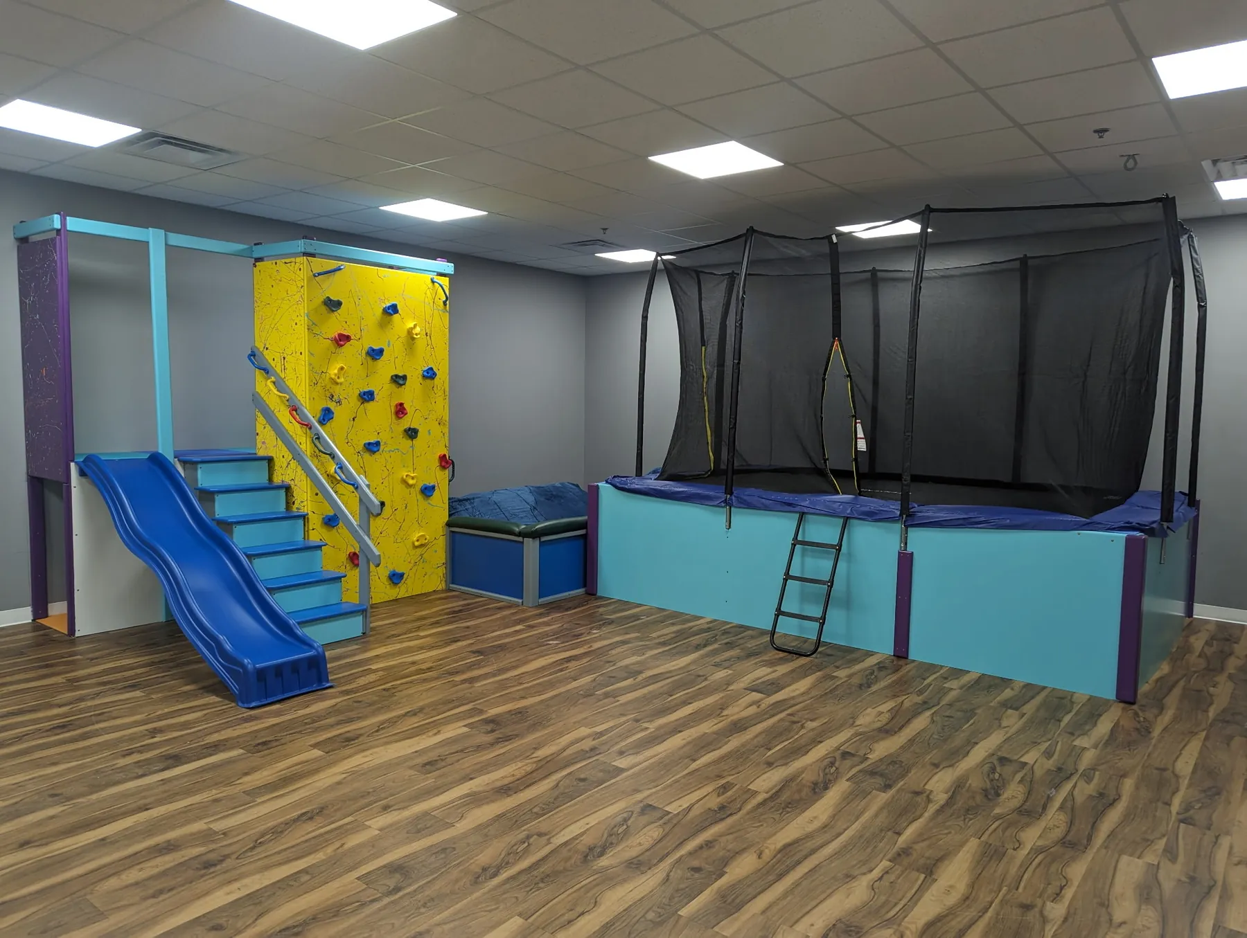 Sensory room with a yellow climbing wall, blue slide and stairs, and a netted trampoline platform on a wood floor
